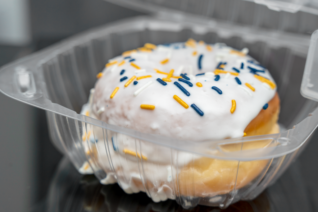 A glazed donut with white icing and blue and gold sprinkles sits in an open plastic container.