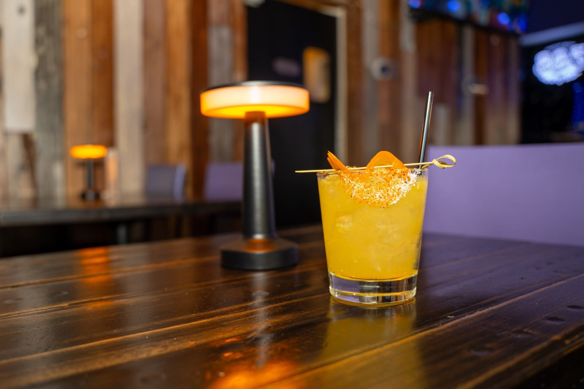 A vibrant orange cocktail garnished with a twist of orange peel sits on a dark wooden table, surroun…